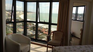 Une chambre d’hôtel dotée d’une grande baie vitrée donnant sur une plage et sur le paysage urbain.