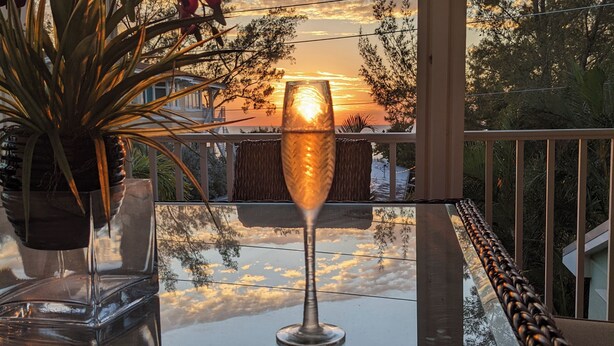 A balcony with a glass table, a glass vase, and a glass with a reflection of the sunset. - Lux Ocean View 4x4 for the Holidays! (Holmes Beach)