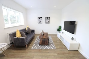A modern living room with a grey sofa, a wooden coffee table, a white entertainment unit, and a large flat-screen TV.