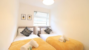 Habitación de hotel con dos camas, cada una con una manta amarilla y una almohada, una ventana con persianas, una lámpara de pared y dos cuadros enmarcados.