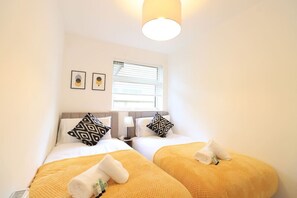 A hotel room with two beds, each with a yellow blanket and a towel. There are two wall-mounted lamps and a window with blinds.