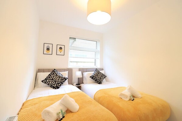 A hotel room with two beds, each with a yellow blanket and a towel. There are two wall-mounted lamps and a window with blinds.