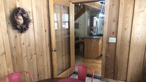 Une salle à manger en bois, avec une table dressée pour quatre personnes, entourée de chaises. Une guirlande décorative est accrochée au mur, et on aperçoit un coin cuisine à travers une porte ouverte.
