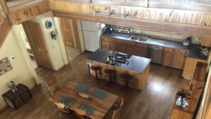 Une cuisine spacieuse dotée de meubles en bois, d’un îlot central avec des tabourets, d’une table à manger avec des chaises et d’un plafond haut avec des poutres apparentes.