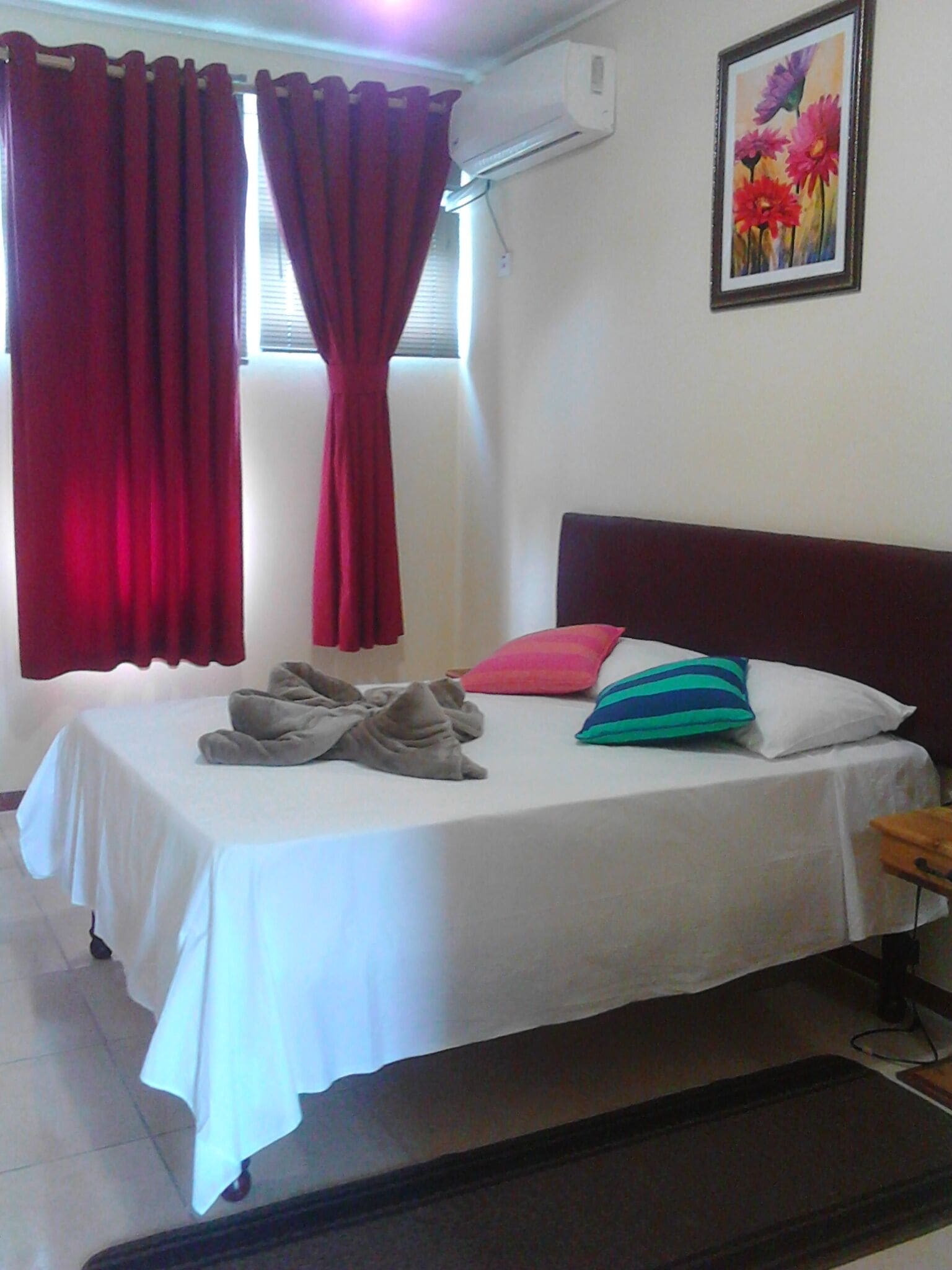 Habitación de hotel con dos camas, cortinas rojas, un cuadro de flores y un equipo de aire acondicionado.