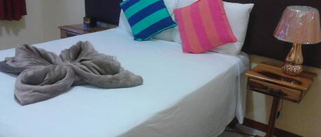 Ein ordentlich gemachtes Bett mit weißer Bettdecke, einem grauen Bademantel und zwei Kissen (eines gestreift blau und grün, eines rosa und weiß). Ein Nachttisch aus Holz ist mit einer Lampe und einer kleinen Schublade ausgestattet.