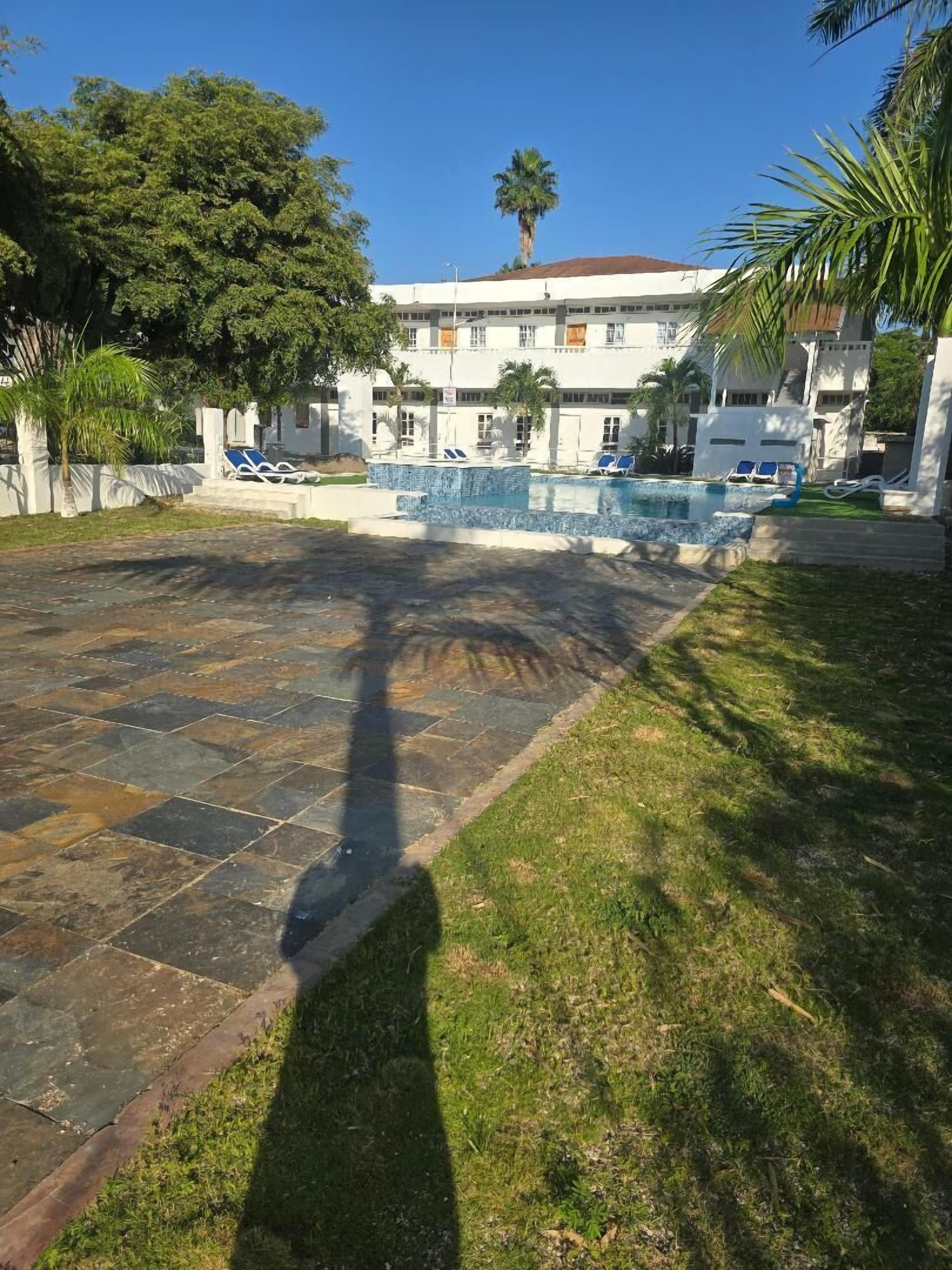 Una zona de piscina con sillones de descanso, un edificio de varios pisos y palmeras.