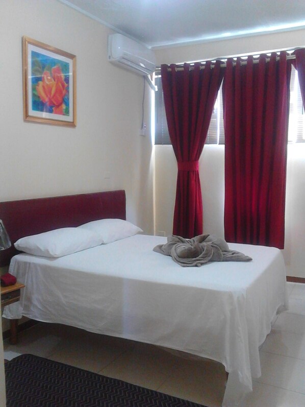 A hotel room with a bed, a nightstand, a lamp, a painting on the wall, and red curtains.