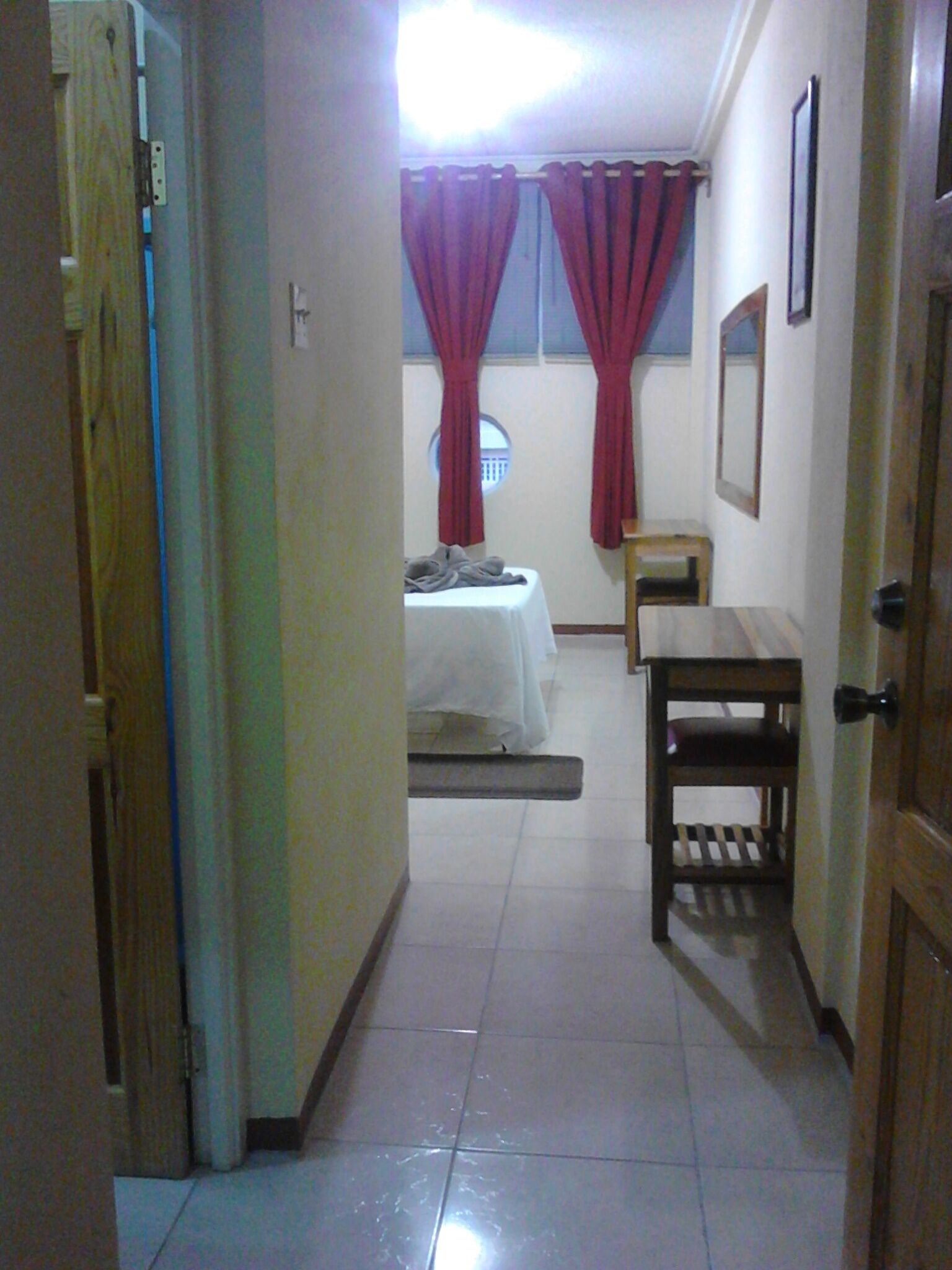 A narrow hallway leading to a bedroom with a bed, a small table, and a window with red curtains.