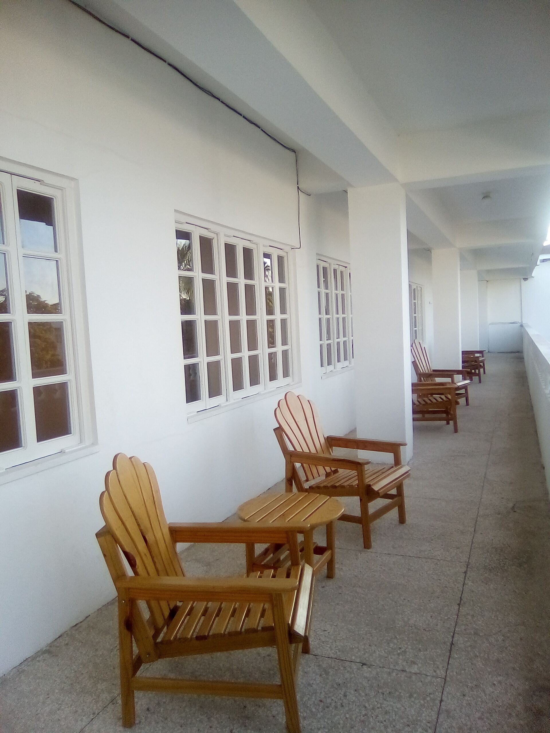 Un pasillo con sillas y mesas de madera, paredes blancas y ventanas grandes.