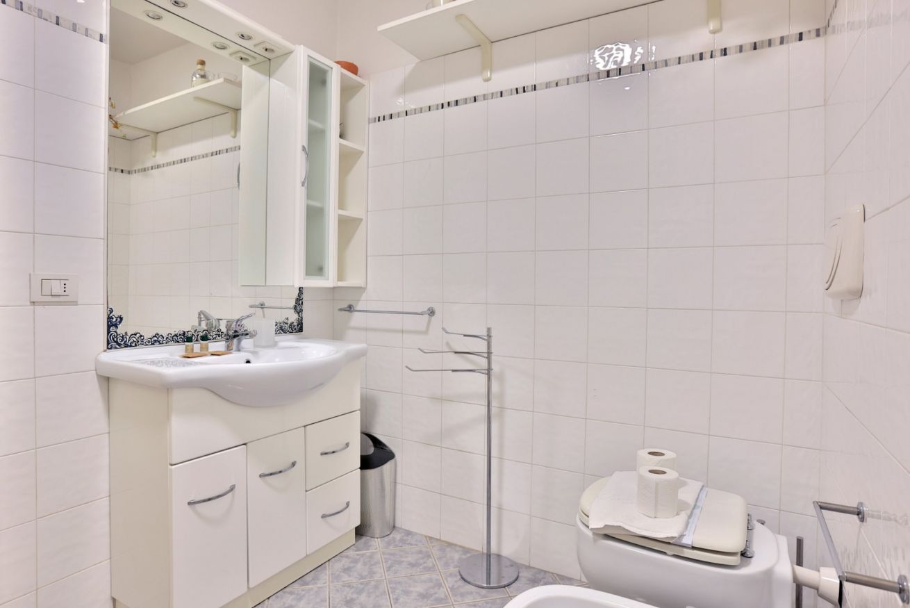 Une salle de bain avec un lavabo blanc, un miroir et des toilettes. On y trouve également un porte-serviettes et une poubelle.