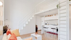 Ein modernes Wohnzimmer mit einem weißen Sofa, einem kleinen Couchtisch und einer Treppe, die in einen Küchenbereich mit weißen Schränken und Barhocker führt.