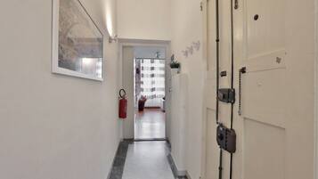 A hallway with a fire extinguisher, a door with a chain, and a view into a room with a bed and a window.