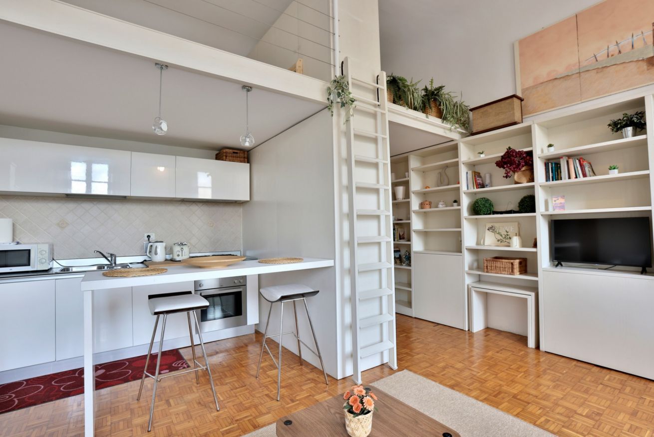 Een moderne keuken met een wit aanrecht, barstoelen, een ladder en een boekenplank met diverse spullen.