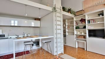 Een moderne keuken met een wit aanrecht, barstoelen, een ladder en een boekenplank met diverse spullen.