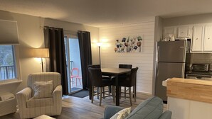 Un espace de vie compact comprenant un canapé, une table à manger avec des chaises, un coin cuisine avec un réfrigérateur et un grille-pain, ainsi qu’une fenêtre avec des stores.