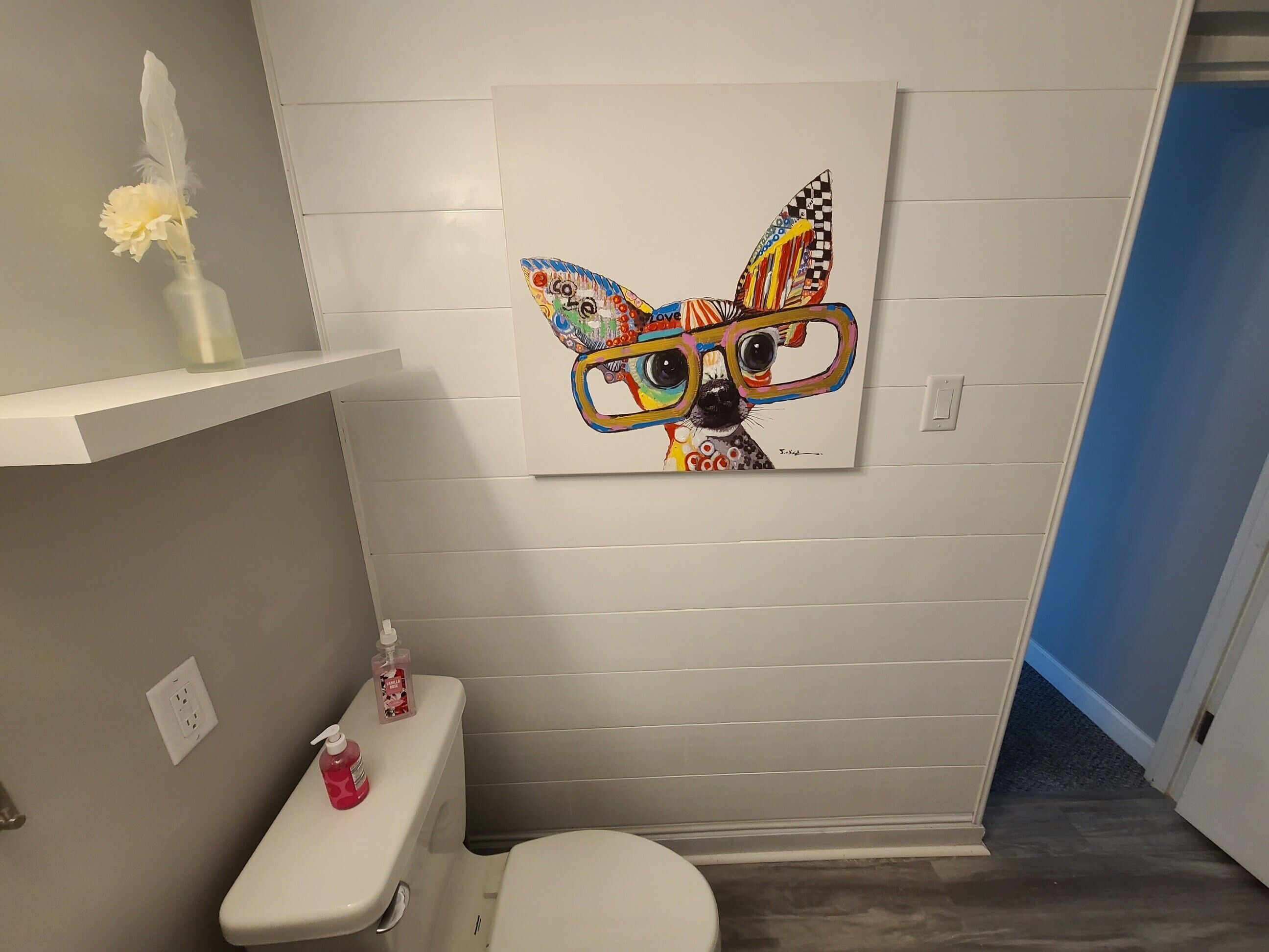 Ein Badezimmer mit einer weißen Toilette, einem Regal mit einer weißen Vase und einer Feder, sowie einem farbenfrohen Gemälde an der Wand.
