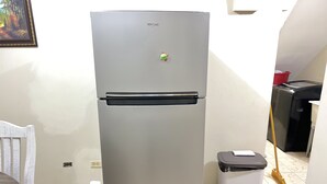 Un refrigerador compacto con pantalla digital, un cubo de basura y una escoba en una habitación con piso de baldosas y una silla blanca.
