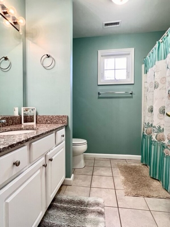 A bathroom with a toilet, a sink with a granite countertop, a mirror, and a shower curtain.