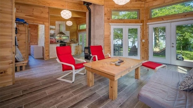 A wooden cabin interior with a dining table, red chairs, a sofa, and large windows.