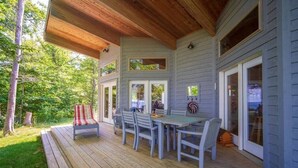 A covered deck with a table and chairs, wooden ceiling, and a view of the surrounding greenery.