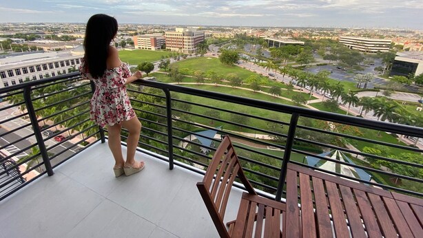 Un balcón con vistas a un paisaje urbano, una mujer de pie y una mesa de madera.