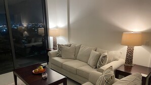 Una sala de estar moderna con un sofá beige, dos mesitas de noche con lámparas, una mesa de centro con fruta, y un ventanal con vistas a la ciudad.