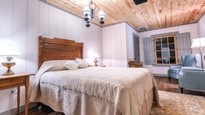 A bedroom with a wooden headboard bed, a nightstand with a lamp, a blue armchair, and a window with shutters. - Bed & Breakfast in a 1795 Log Cabin on working farm, George Washington, Sr Suite (Swoope)