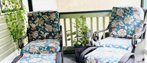 A covered porch with two wicker chairs and a small table, surrounded by potted plants and a wooden railing.