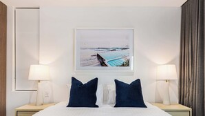 A hotel room with a bed, two bedside lamps, a framed picture of a beach, and a window with curtains. - The Arden 7 Coogee New Beachside Ocean View Apt (Coogee)