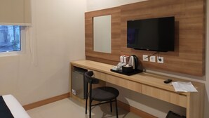 Habitación de hotel con escritorio de madera, refrigerador pequeño, silla, televisor montado en la pared y ventana con vista a edificios.