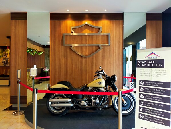 A motorcycle in a showroom with a sign advising to stay safe and healthy, and a wooden wall with a logo. - Sukho Hotel (Ciakar)