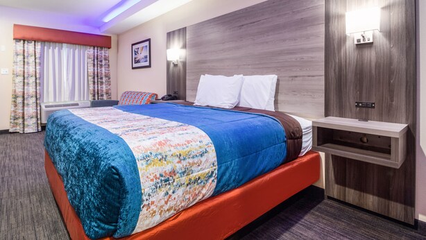 A hotel room with a large bed, a nightstand, a wall-mounted lamp, and a window with curtains. - Scottish Inn & Suites Houston Willowbrook (Houston)