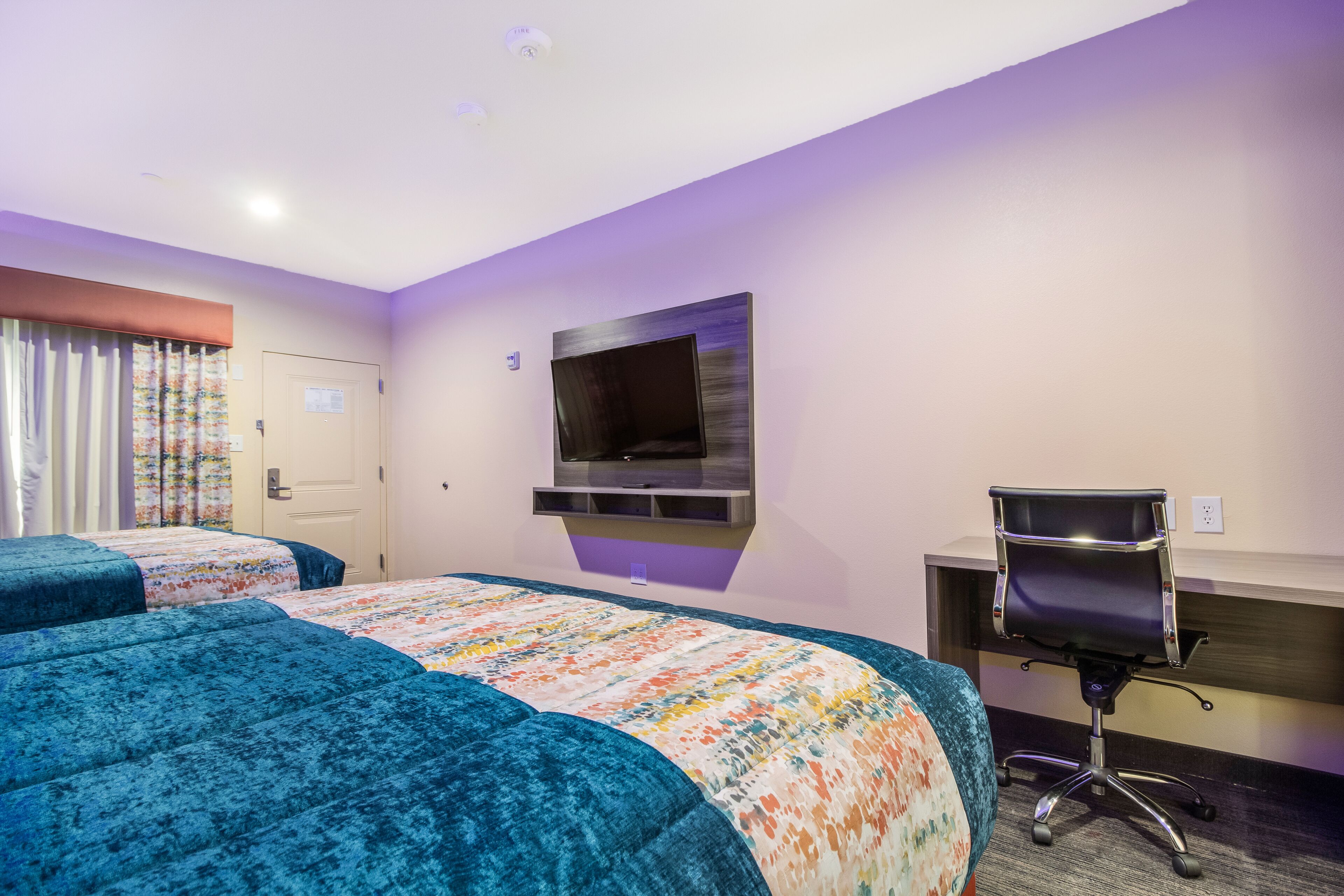 A hotel room with two beds, a desk, a chair, a television, and a door.