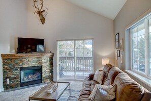A living room with a stone fireplace, a television mounted above it, a leather sofa, and a large window providing a view of the outside. - Weekly Deals! Remodeled Retreat + Hot Tub + Garage (Steamboat Springs)
