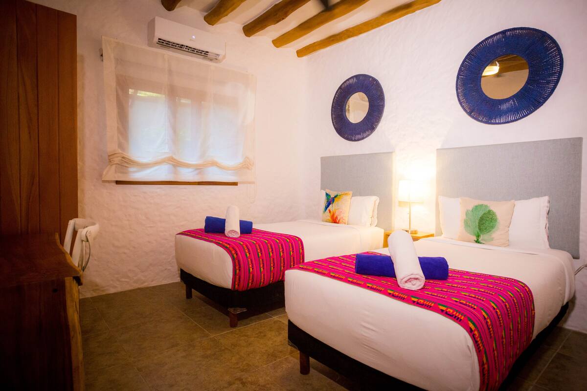 A hotel room with two beds, each with a white mattress and a colorful blanket. There are two round mirrors on the wall, a window with sheer curtains, and a wooden ceiling.