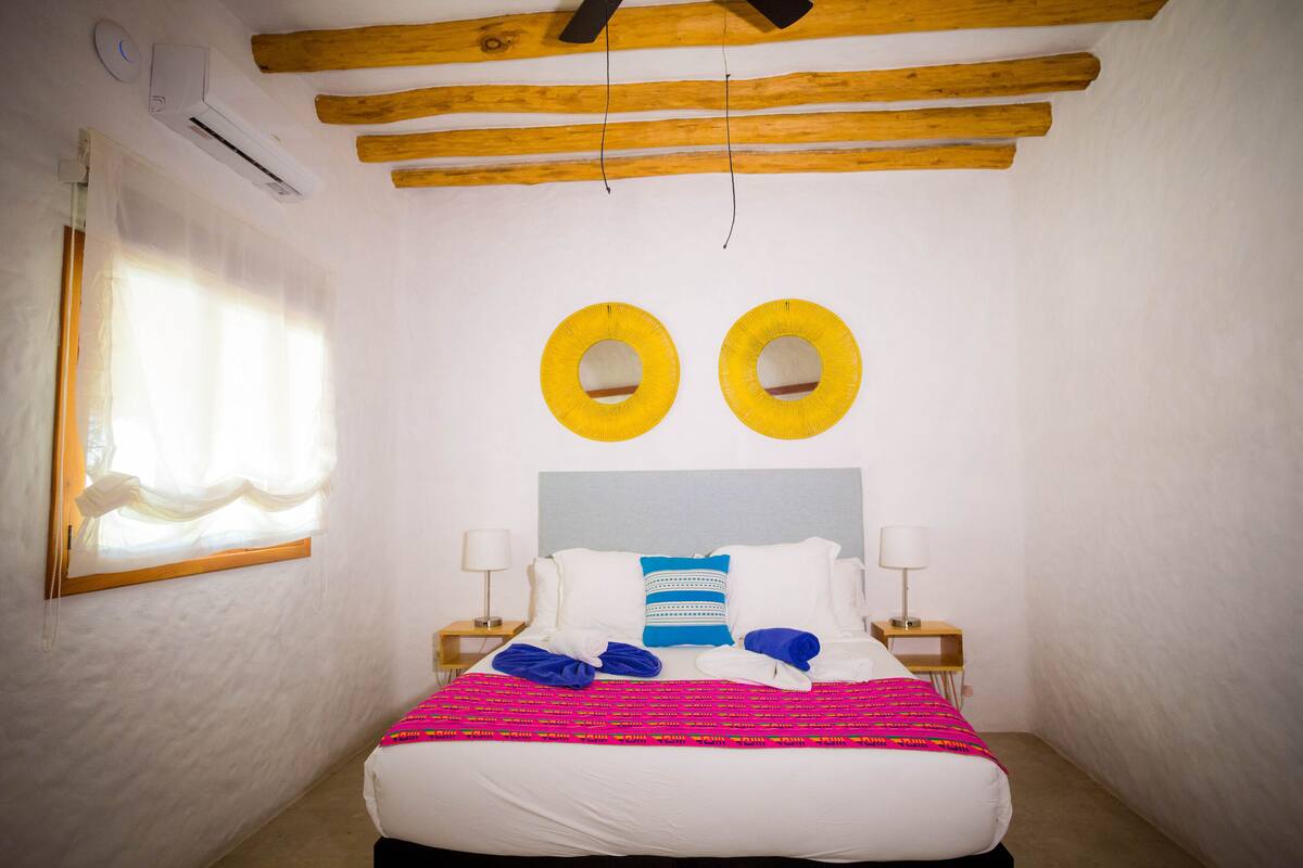 A hotel room with a bed, two bedside tables, a window with curtains, and a ceiling with exposed beams.