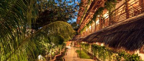 A pathway leading to a building with a thatched roof and wooden lattice walls, surrounded by potted plants and palm trees.