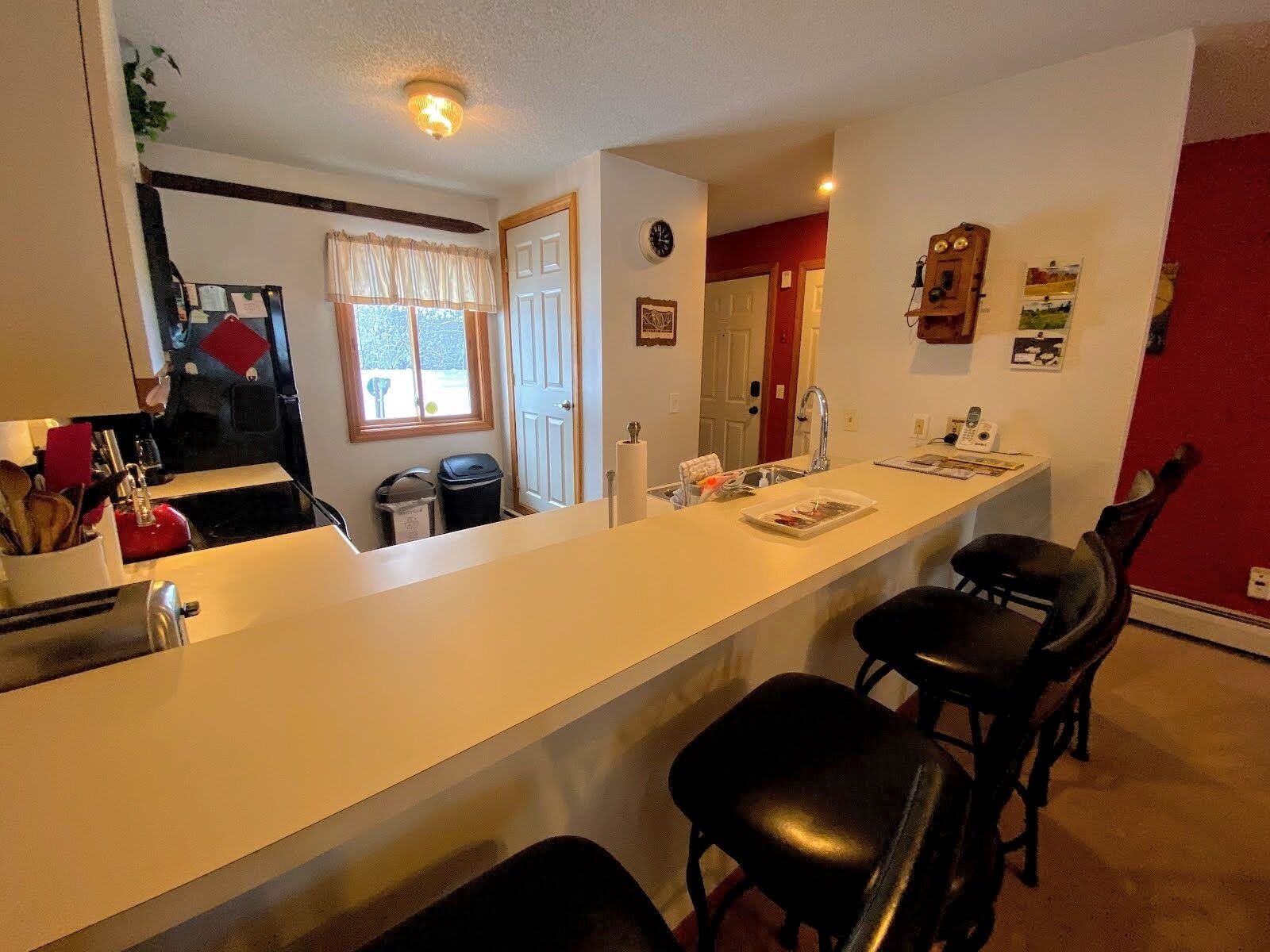 Een keuken met een wit aanrecht, een spoelbak en een raam met gordijnen. Er staan twee zwarte stoelen en is een rode muur.