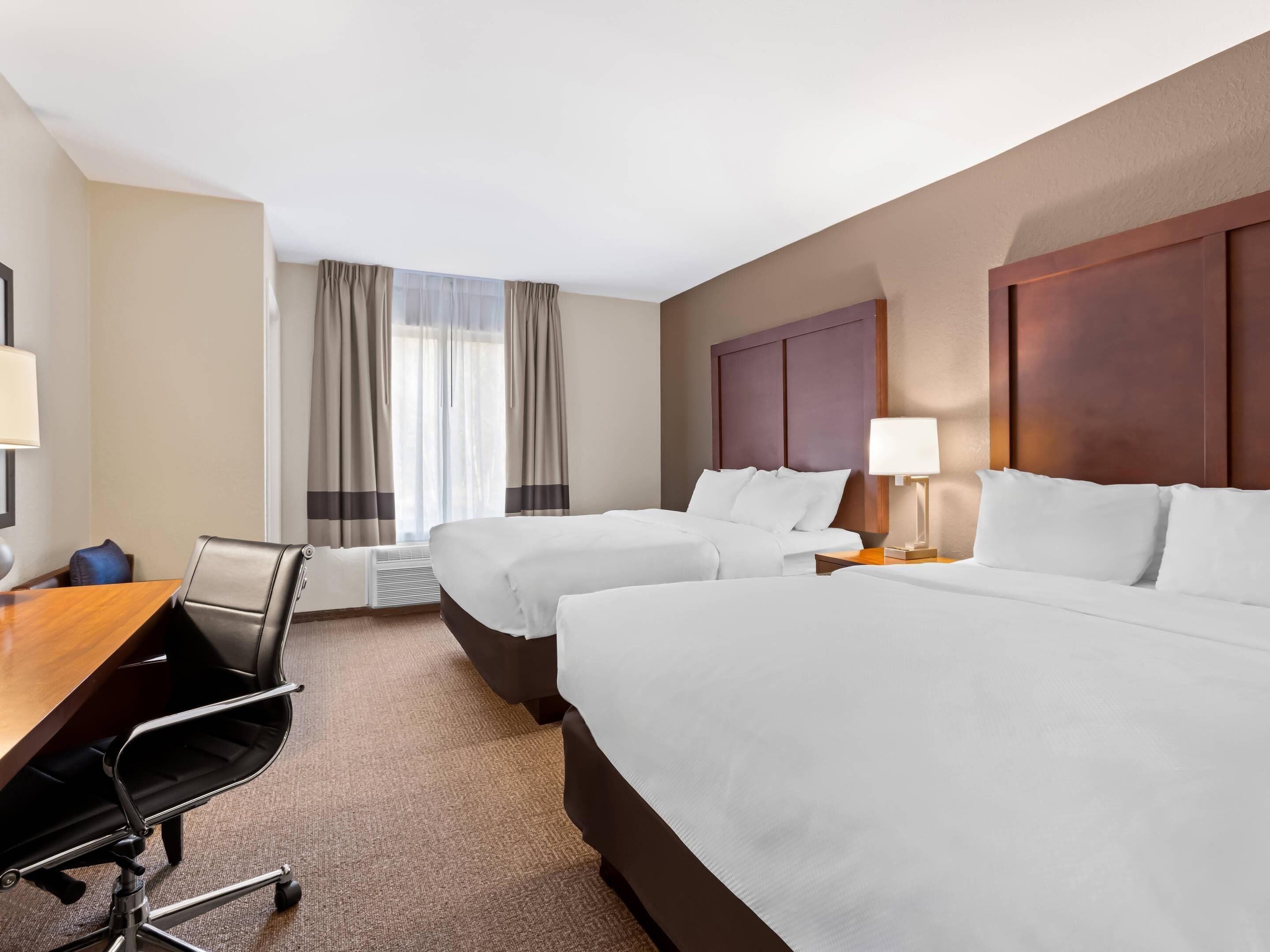 A hotel room with two beds, a desk, a chair, and a window with curtains.
