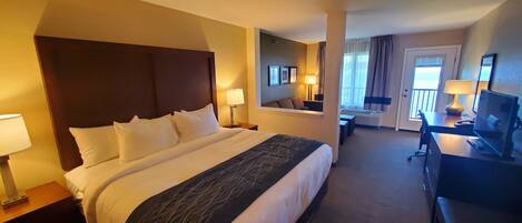 A hotel room with a large bed, a desk with a computer, a chair, and a TV. There is a door leading to a balcony with a view.
