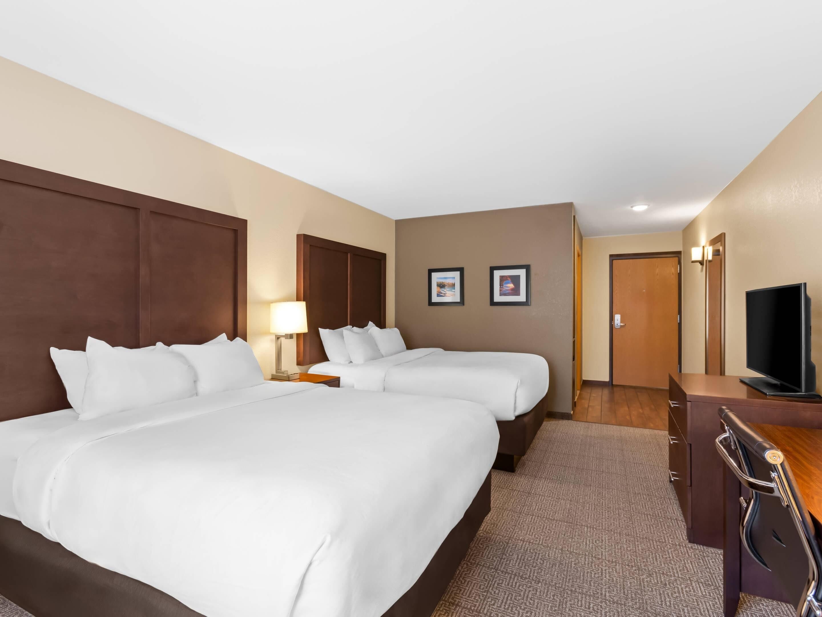A hotel room with two beds, a desk, a television, and framed pictures on the wall.