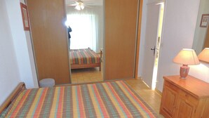 A bedroom with a bed, a bedside table with a lamp, a wardrobe, and a mirror. - 3-Star F2 w/ Parking, Terrace, & Thau Lagoon View near Thermal Baths (Balaruc-les-Bains)