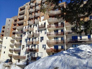 Um edifício residencial de vários andares com varandas, uma pista de esqui em primeiro plano e um céu azul límpido.