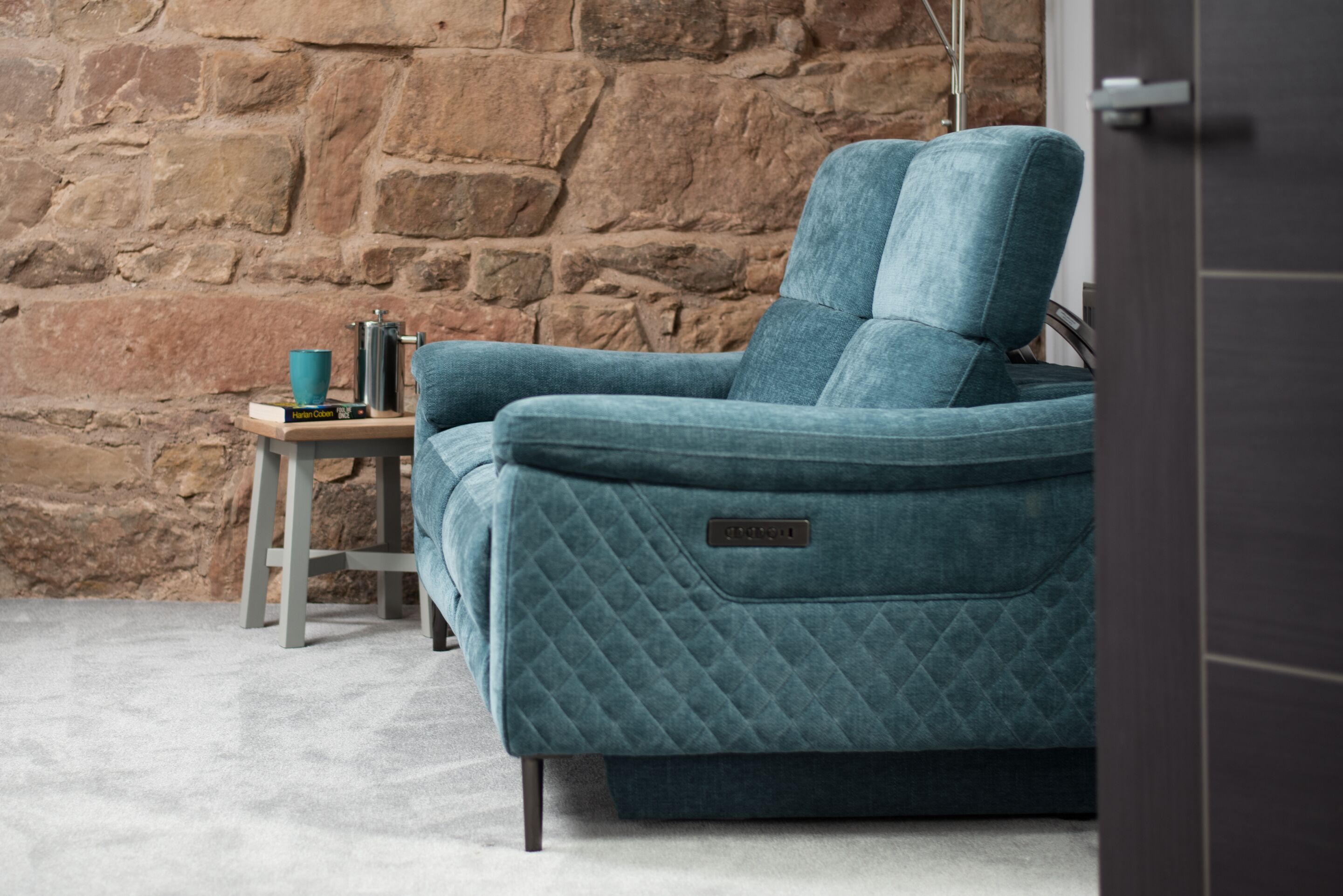 A teal upholstered chair with a quilted pattern, a small wooden side table with a blue mug and books, and a stone wall in the background.
