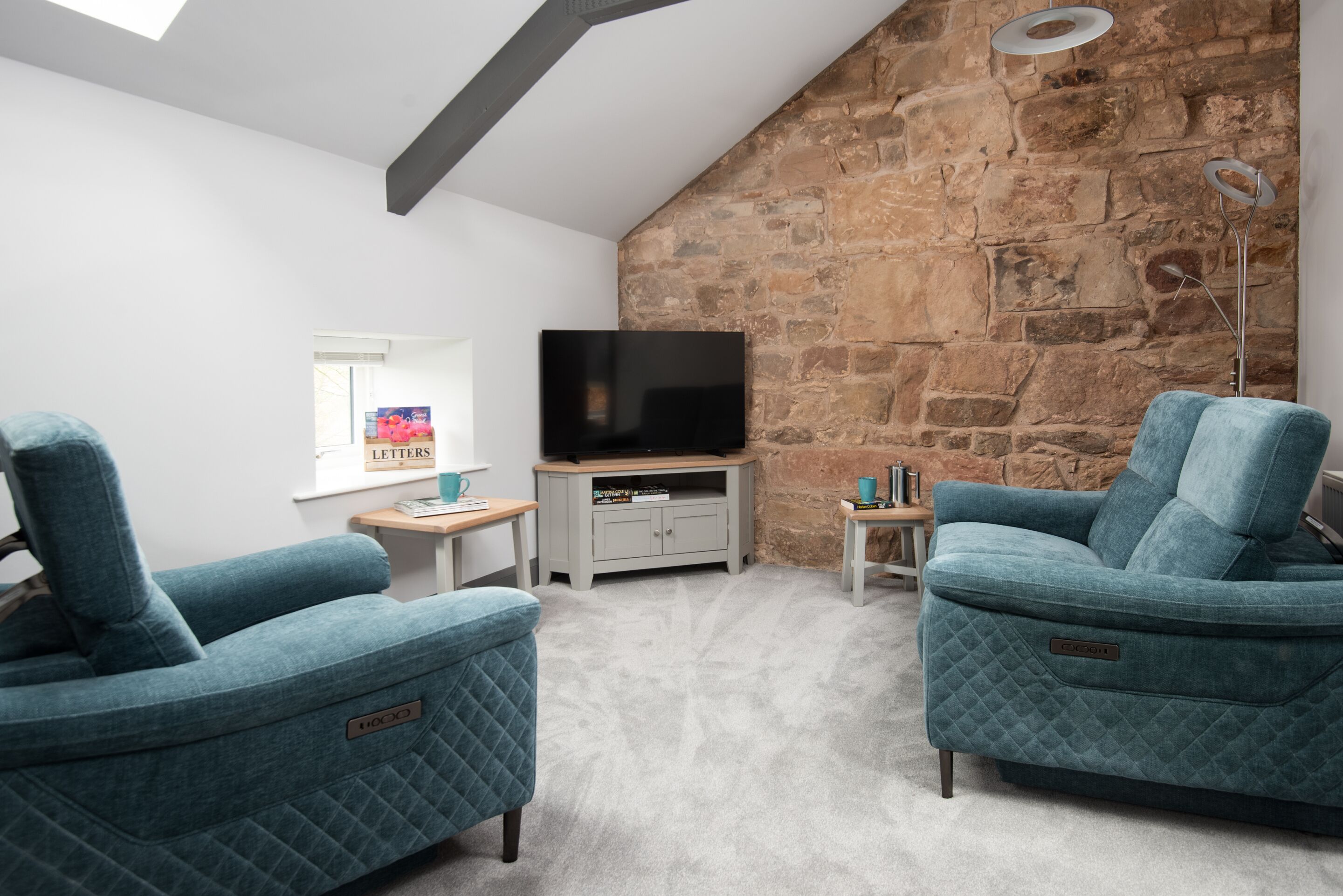 A cozy living room with two blue armchairs, a flat-screen TV on a wooden stand, a stone accent wall, and a small side table with a lamp.