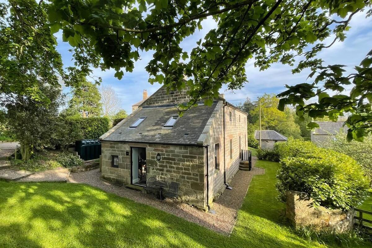 Une petite maison en pierre avec un toit en pente, un petit porche et un jardin comprenant une pelouse et des arbres.