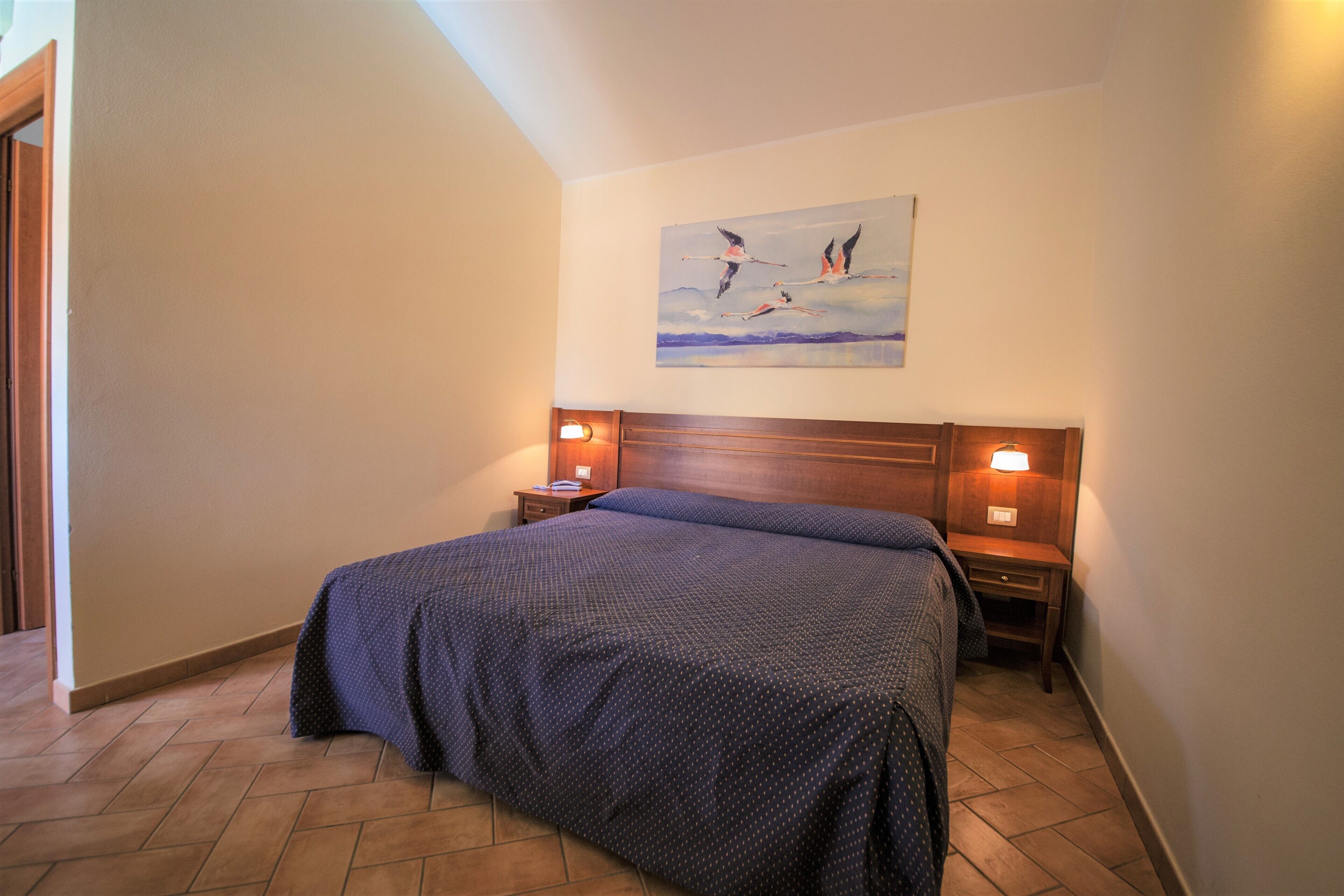 A bedroom with a large bed, two bedside tables with lamps, a wooden headboard, and a painting on the wall.
