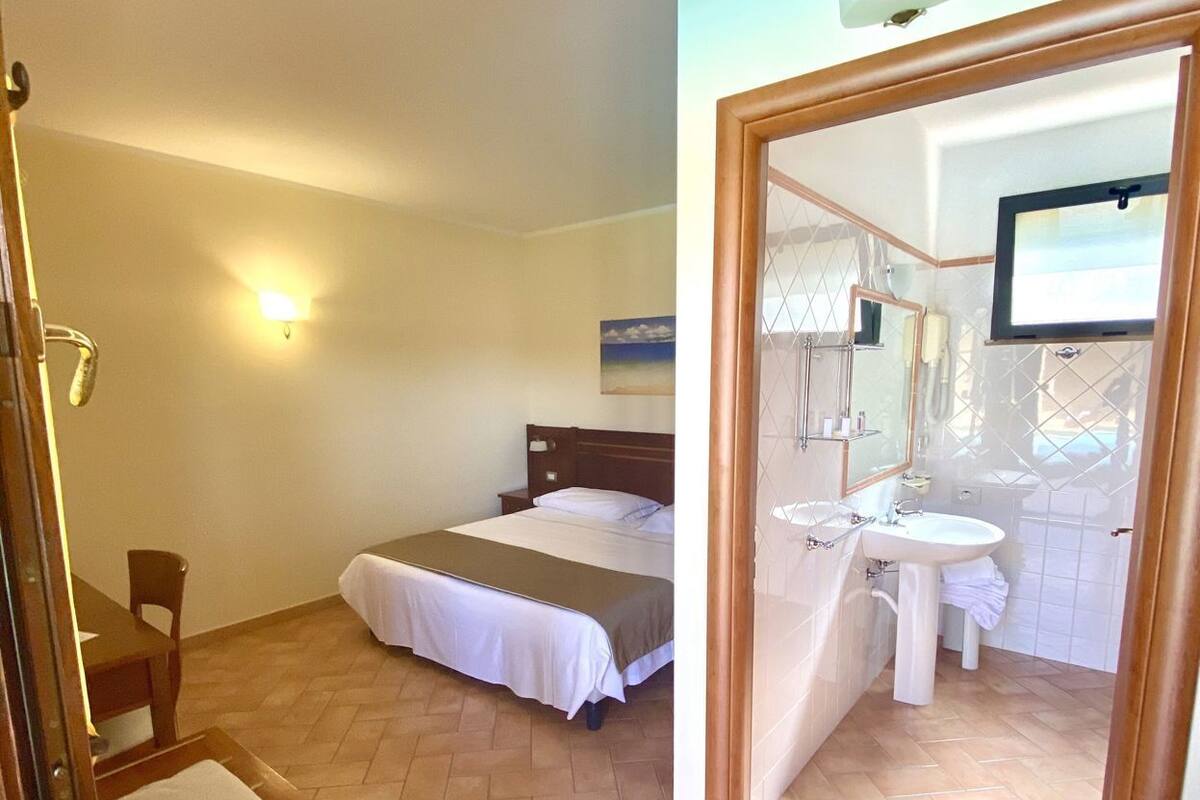 Une chambre à coucher comprenant un lit, une table de chevet, une chaise et un bureau avec une autre chaise. Il y a aussi une salle de bain avec un lavabo, un miroir et un porte-serviettes.