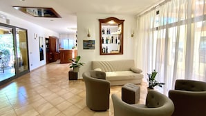 Ein großzügiger Wohnbereich mit einer Couch, Sesseln und einem Couchtisch. Im Hintergrund befindet sich eine Küchenzeile, ein Esstisch und ein großes Fenster mit Vorhängen.